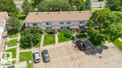 Multi-unit residential building with light-colored siding, brown roofing, individual front entrances, and an adjacent paved parking area - 