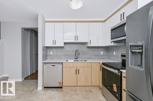 Modern kitchen featuring two-tone cabinetry with white upper and wood-finish lower cabinets, stainless steel appliances, light-colored countertops, and a full-height tile backsplash - 24 14511 52 Street, Edmonton, AB 