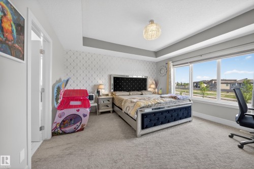 Spacious bedroom featuring large windows, tray ceiling with a contemporary chandelier, textured accent wall, and light gray carpeting - 1927 18 Street, Edmonton, AB 