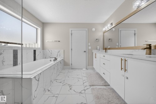 Spacious bathroom featuring a dual vanity with white cabinetry and gold-toned hardware, a large framed mirror, and an enclosed bathtub with marble-look tile surround - 1927 18 Street, Edmonton, AB 