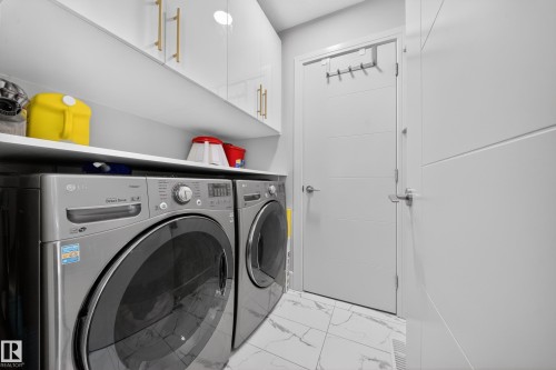 Dedicated laundry area featuring a front-loading washer and dryer, white cabinetry with gold-finish hardware, a white utility shelf, and white tile flooring - 1927 18 Street, Edmonton, AB 