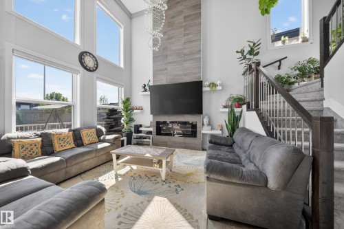 Two-story living area featuring a tile-clad fireplace, high ceilings, abundant natural light from large windows, a carpeted staircase with wood and metal railing, and wood-finish flooring - 1927 18 Street, Edmonton, AB 