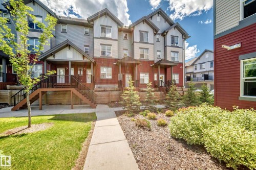 Multi-level townhome featuring a covered entrance porch, exterior stairs with dark railings, and a landscaped front yard with grass and plantings - 118 655 Watt Boulevard Sw, Edmonton, AB 