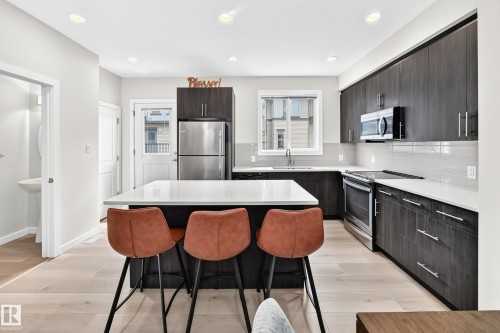 Contemporary kitchen featuring a central island with a white countertop, dark wood-finish cabinetry, stainless steel appliances, light wood-finish flooring, and a white subway tile backsplash - 118 655 Watt Boulevard Sw, Edmonton, AB 