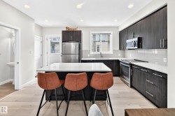 Contemporary kitchen featuring a central island with a white countertop, dark wood-finish cabinetry, stainless steel appliances, light wood-finish flooring, and a white subway tile backsplash - 