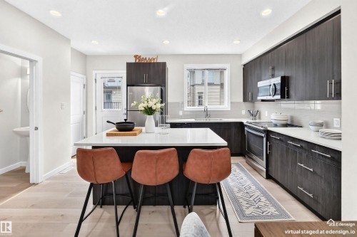 Modern kitchen featuring a central island with a light-toned countertop, dark wood-finish cabinetry, stainless steel appliances, a subway tile backsplash, and light wood-finish flooring - 118 655 Watt Boulevard Sw, Edmonton, AB 