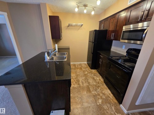 Kitchen featuring dark wood-finish cabinetry, black countertops, and a double basin stainless steel sink - 319 5816 Mullen Place, Edmonton, AB 