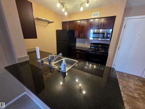 Kitchen featuring a dark granite countertop with a double-basin sink, rich wood-finish cabinetry, and black appliances including a refrigerator and range - 319 5816 Mullen Place, Edmonton, AB 