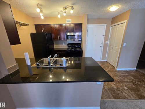 Kitchen featuring dark wood-finish cabinetry, black appliances, a double basin stainless steel sink, and a black granite-look countertop island - 319 5816 Mullen Place, Edmonton, AB 