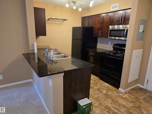 Compact kitchen featuring dark wood-finish cabinetry, black appliances, a double basin stainless steel sink, and a speckled dark countertop - 319 5816 Mullen Place, Edmonton, AB 