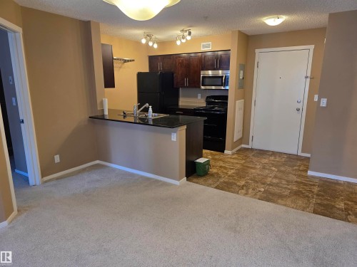 Open-concept living area featuring a kitchen with dark cabinetry, stainless steel microwave, black appliances, and a breakfast bar with a dark countertop - 319 5816 Mullen Place, Edmonton, AB 