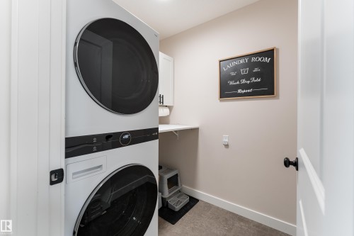 Dedicated laundry area featuring stacked front-load appliances, a white wall-mounted shelf, and a white storage cabinet with black hardware - 584 Meadowview Drive, Fort Saskatchewan, AB 