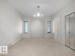 Spacious room featuring neutral carpeting, white walls, two vertical windows with blinds, a modern track light fixture, and mirrored closet doors - 