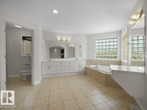 Spacious bathroom featuring a dual vanity with white cabinetry and an arched mirror, a built-in soaking tub with tiled surround, and glass block windows - 2423 Tegler Green, Edmonton, AB 