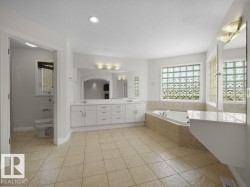 Spacious bathroom featuring a dual vanity with white cabinetry and an arched mirror, a built-in soaking tub with tiled surround, and glass block windows - 