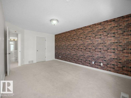 Spacious room featuring a prominent brick-patterned accent wall, light-toned carpet flooring, a ceiling-mounted light fixture, and white painted walls - 2423 Tegler Green, Edmonton, AB 