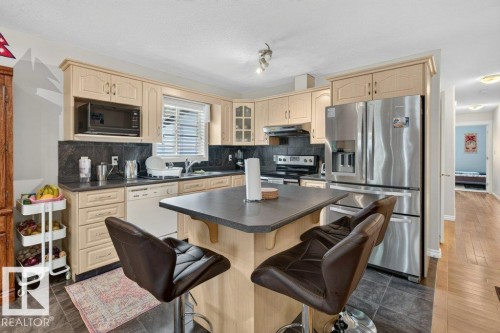 Kitchen featuring light wood cabinetry, a central island with countertop seating, stainless steel appliances, dark stone-look countertops, and tile flooring - 11330 101 Street, Edmonton, AB 