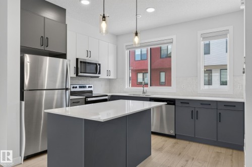 Modern kitchen featuring a central island with a light-toned countertop, stainless steel appliances, two-tone cabinetry, wood-finish flooring, and pendant lighting - 33 11910 35 Avenue, Edmonton, AB 