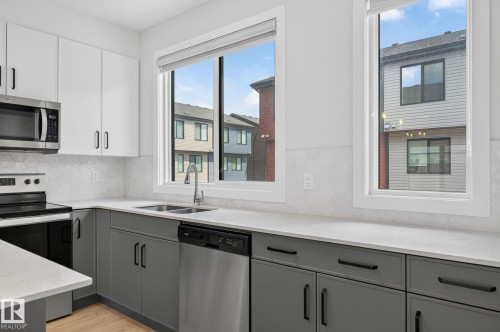 Contemporary kitchen featuring two-tone cabinetry, stainless steel appliances, a double basin sink, light-toned countertops, and a hexagonal tile backsplash - 33 11910 35 Avenue, Edmonton, AB 