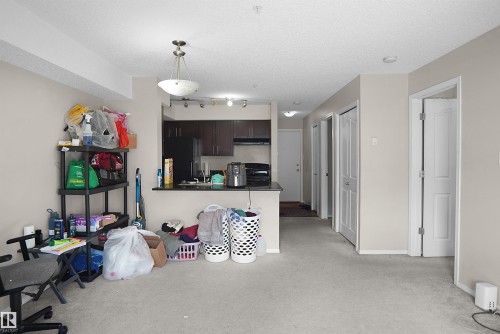Open-concept living space featuring neutral-toned walls, light-colored carpeting, and a kitchen area with dark cabinetry, a black refrigerator, and a dark countertop - 215 1070 Mcconachie Blvd, Edmonton, AB 