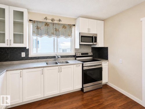 Kitchen featuring white cabinetry, stainless steel appliances, a double basin sink, dark mosaic tile backsplash, and wood-finish flooring - 4304 121 Avenue Nw, Edmonton, AB - Indoor Photo Showing Kitchen With Double Sink