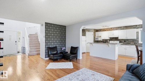 Open-concept living area featuring wood-finish flooring, a contemporary accent wall, and a bright kitchen with white cabinetry and a stone-finish backsplash - 3591 Mclean Crescent, Edmonton, AB 