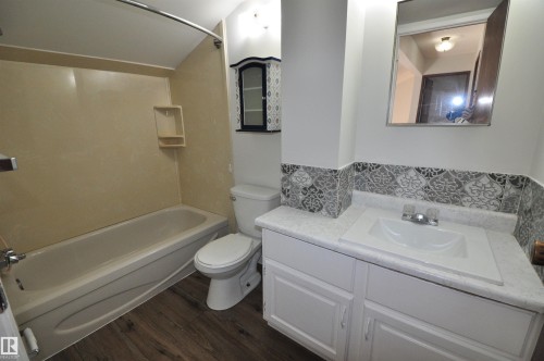 Bathroom featuring a single vanity with an integrated sink, patterned tile backsplash, wood-finish flooring, a toilet, and a combination tub and shower with a beige surround - 617 Millbourne Road E, Edmonton, AB 