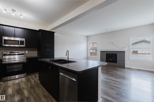 Open-concept kitchen and living area featuring dark cabinetry, stainless steel appliances, a kitchen island with a double basin sink, wood-finish flooring, and a fireplace with a tile surround - 636 Mcdonough Link Link, Edmonton, AB 