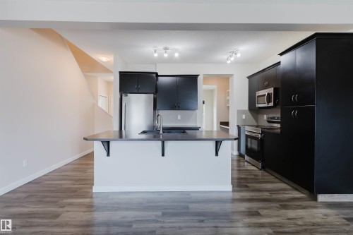 Open-concept kitchen featuring an island with seating, dark shaker-style cabinetry, stainless steel appliances, and wood-finish flooring - 636 Mcdonough Link Link, Edmonton, AB 