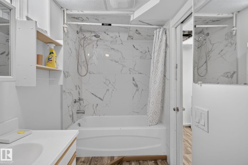 Bathtub with white marble-look surround and chrome fixtures, complemented by wood-finish flooring and a white vanity with an integrated sink - 12238 48 Street, Edmonton, AB 