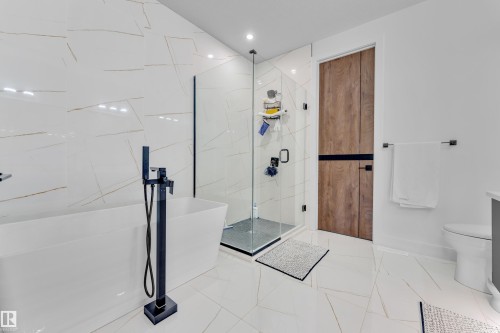 Contemporary bathroom featuring a freestanding soaking tub with a matte black floor-mounted faucet, a glass-enclosed shower with a dark framed door, and large format white tile flooring extending up the shower wall - 16816 49 Street, Edmonton, AB 
