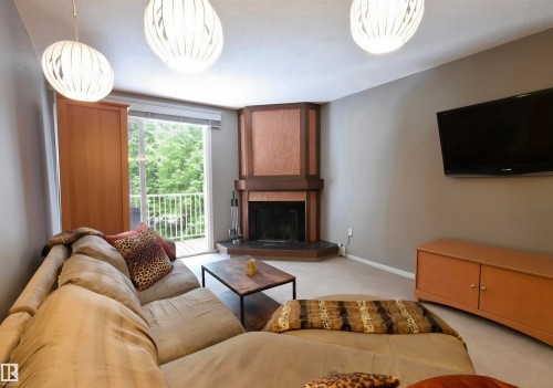 Living area featuring a corner fireplace with a textured surround, a sliding glass door leading to a balcony with white railings, and three contemporary pendant light fixtures - 302 10335 118 Street, Edmonton, AB 