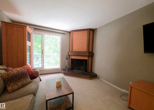 Living area featuring a corner fireplace with a wood surround and textured paneling, light-colored carpeting, and a sliding glass door opening to a balcony - 302 10335 118 Street, Edmonton, AB 