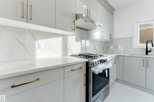 Modern kitchen featuring white cabinetry with gold-tone hardware, white countertops, and a full backsplash with subtle veining - 1536 64 Ave, Rural Leduc County, AB 