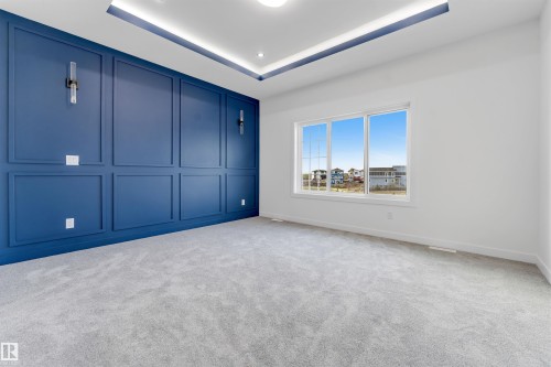 Spacious room featuring a deep blue accent wall with decorative paneling and integrated sconces, soft gray carpeting, a large white-framed window, and a tray ceiling with recessed lighting - 1536 64 Ave, Rural Leduc County, AB 