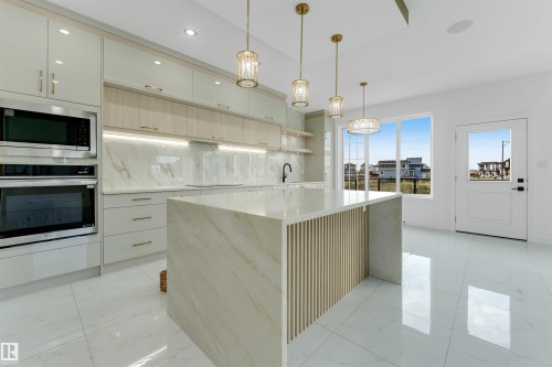 Modern kitchen featuring a waterfall island with slatted wood detailing, integrated stainless steel appliances, marble-effect slab backsplash, glossy white cabinetry, and polished tile flooring - 1536 64 Ave, Rural Leduc County, AB 