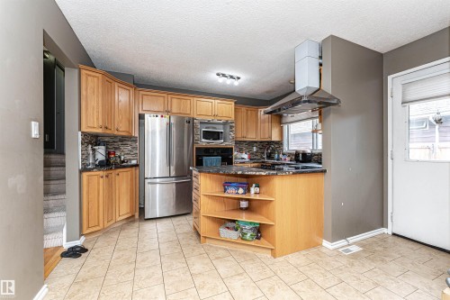 Kitchen featuring warm wood cabinetry, a central island with open shelving, stainless steel appliances, a mosaic tile backsplash, and light-toned tile flooring - 11512 24 Avenue, Edmonton, AB 