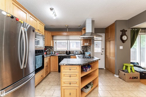 Kitchen featuring wood cabinetry, stainless steel refrigerator, tile backsplash, and a central island with a cooktop - 11512 24 Avenue, Edmonton, AB 