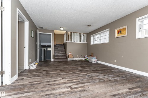 Spacious room featuring wood-finish flooring, neutral-toned walls, multiple windows, and a staircase with white railings - 11512 24 Avenue, Edmonton, AB 