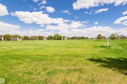 Expansive green space featuring a large grassy field, distant residential structures, and mature trees under a bright sky - 