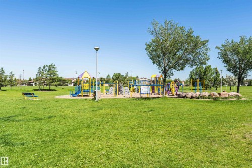 Expansive green space featuring a colorful playground structure with slides and climbing elements, surrounded by mature trees, a sand surface, a park bench, and a modern streetlamp - 11512 24 Avenue, Edmonton, AB 