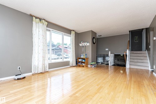 Bright living area featuring light wood-finish flooring, large picture windows, neutral wall tones, and a carpeted staircase with a white railing - 11512 24 Avenue, Edmonton, AB 