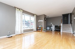 Bright living area featuring light wood-finish flooring, large picture windows, neutral wall tones, and a carpeted staircase with a white railing - 