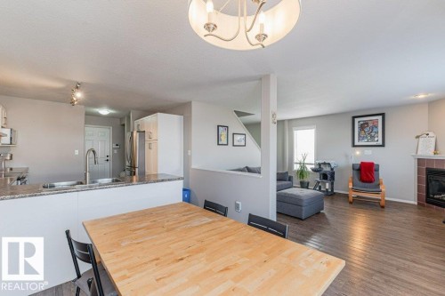 Open-concept living space featuring a dining area with a light wood-finish table, a kitchen island with a stone-finish countertop, and a living room with wood-finish flooring and a fireplace with a brick surround - 1816 36 Avenue, Edmonton, AB 