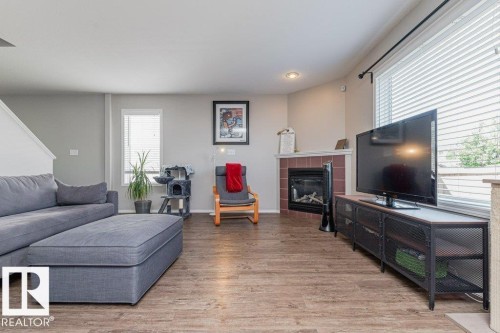Wood-finish flooring throughout, featuring a corner fireplace with a red tile surround, large windows, recessed lighting, and a partial staircase visible - 1816 36 Avenue, Edmonton, AB 