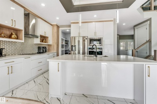 Modern kitchen featuring a large central island with a built-in sink, white cabinetry with gold-finish hardware, stainless steel appliances, a hexagonal tile backsplash, and marble-look tile flooring - 715 Ebbers Place Nw, Edmonton, AB 