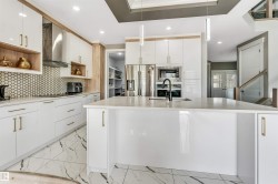 Modern kitchen featuring a large central island with a built-in sink, white cabinetry with gold-finish hardware, stainless steel appliances, a hexagonal tile backsplash, and marble-look tile flooring - 