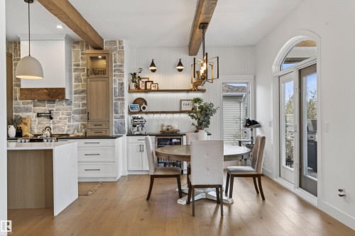 Open-concept kitchen and dining area featuring wood-finish flooring, exposed ceiling beams, stone accent wall, white cabinetry, and an arched glass patio door - 711 Beach Avenue, Cold Lake, AB - Indoor