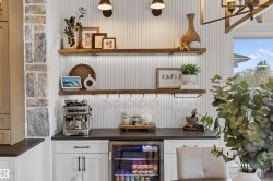 Custom beverage bar featuring white shaker cabinetry, dark countertops, a built-in beverage refrigerator, wood-finish floating shelves with integrated lighting, and vertical plank wall paneling - 