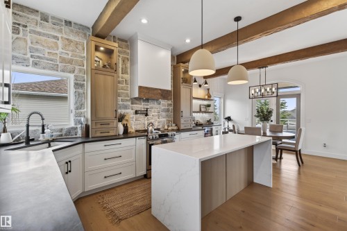 Gourmet kitchen featuring a stone accent wall, wood-finish flooring, exposed ceiling beams, a central island with a waterfall countertop, and built-in cabinetry - 711 Beach Avenue, Cold Lake, AB - Indoor Photo Showing Kitchen With Upgraded Kitchen